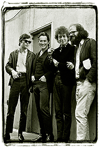 A black and white photograph of four men against the backdrop of a wall and a door. The photo style is relaxed and candid.